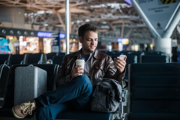 Vacker leende man i fritidskläder hålla bagage och skicka meddelanden genom sin mobiltelefon när du sitter i hallen på flygplatsen med kaffekopp — Stockfoto