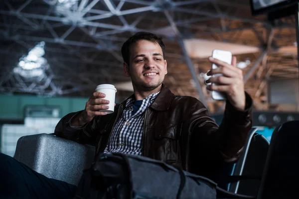 Porträtt av en ung man skrattar och tar selfie med kaffe på flygplatsen väntar på sin flight — Stockfoto