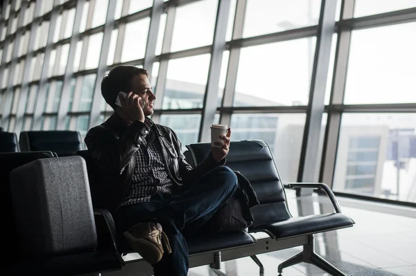 Retrato de un joven guapo vestido con ropa de estilo casual sentado en el banco en el aeropuerto moderno usando un teléfono inteligente. Pasajero viajando con maleta haciendo llamada, mientras espera su vuelo —  Fotos de Stock