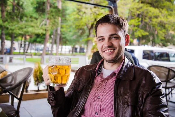 Salud, joven guapo brindando con cerveza y mirando a la cámara sonriendo mientras está sentado en el mostrador del bar — Foto de Stock