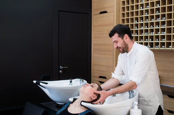 Cabeleireiro bonito lavando o cabelo feminino com concentração. Ele está segurando uma torneira de água. Mulher inclinando a cabeça na pia . — Fotografia de Stock