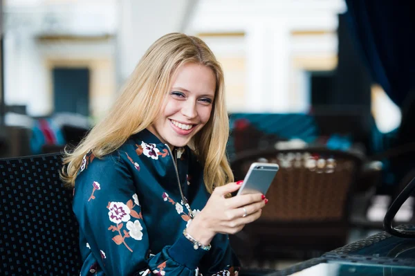 Hermosa joven sentada en la cafetería urbana al aire libre de moda, sonriendo y mensaje de texto en su teléfono inteligente. Horizontal — Foto de Stock