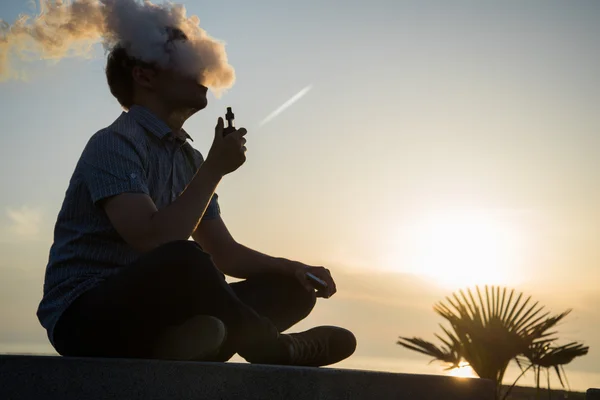 Vaping young man with, produces vapor on sunset sky background at the sea coast promenade, place for text — Stock Photo, Image