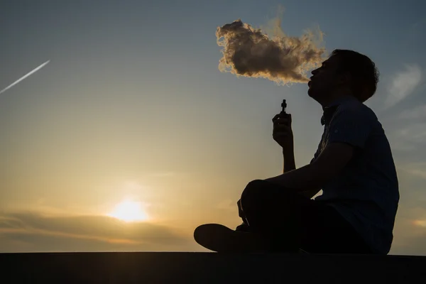 Vaping jovem com, produz vapor no fundo céu por do sol no passeio marítimo costa, lugar para texto — Fotografia de Stock