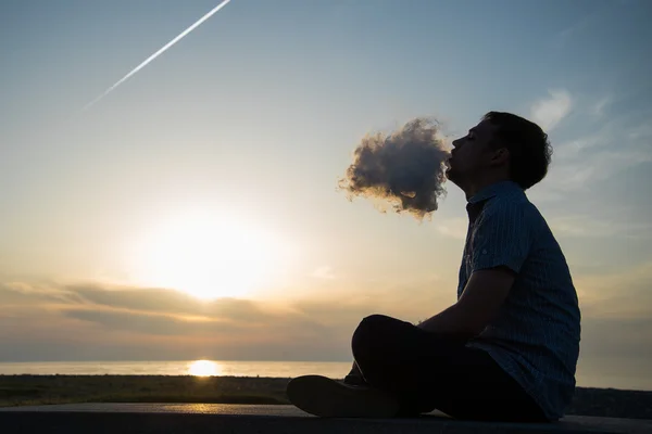 Siluet manusia merokok pipa di topi saat matahari terbenam. Fotografer bertopi bisbol merokok pipa saat matahari terbenam Oleh laut. Perjalanan, petualangan, meditasi, relaksasi, liburan . — Stok Foto