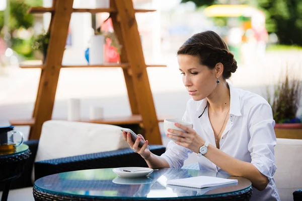 Retrato de una joven empresaria tomando café y usando teléfono móvil mientras está sentada en la cafetería de la acera — Foto de Stock