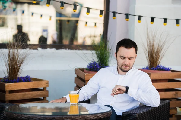 Un hombre de negocios guapo está esperando a un cliente en la cafetería. Está tocando el reloj y mirándolo con anticipación. El hombre está sentado en la mesa al aire libre y sonriendo. Está bebiendo jugo de naranja. — Foto de Stock