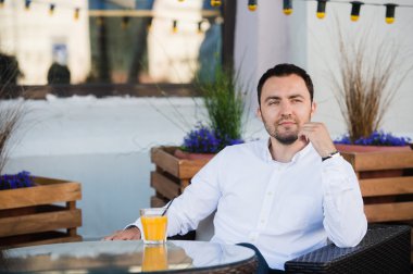 Yakışıklı işadamı kafeteryada müşteri bekliyor. Saate dokunuyor ve beklentiyle bakıyor. Adam dışarıda masada oturuyor ve gülümsüyor. Portakal suyu içiyor.