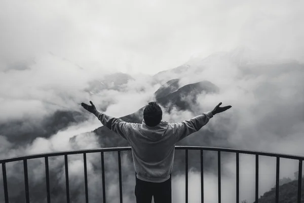 Zwart-wit getinte afbeelding jongeman hoodie dragen op de rand van een klif en schreeuwt. Hij toont zijn handen tegen mistige bergen met dikke wolken zweven naar beneden — Stockfoto