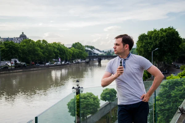 Молодой красивый мужчина с электронной сигаретой vaping и стоя на террасе возле реки в европейском городе — стоковое фото