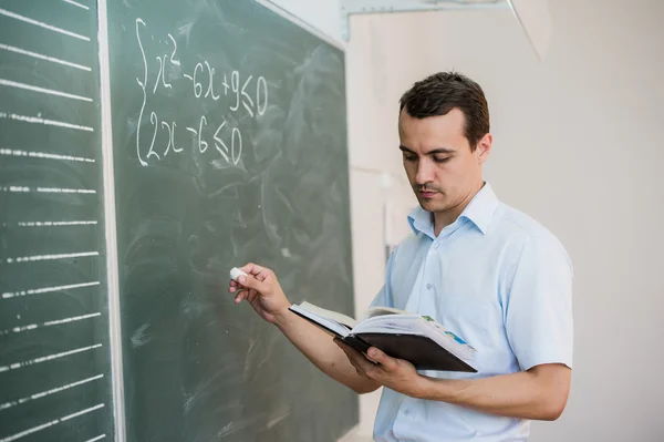 Young male teacher or student holding chalk writing on chalkboard in classroom — ストック写真