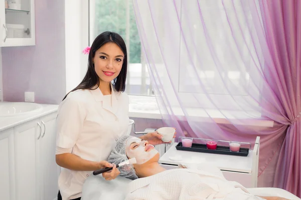 Joven cosmetólogo alegre aplicando máscara en la cara femenina con un cepillo. Ella está de pie y sonriendo. La mujer que miente — Foto de Stock