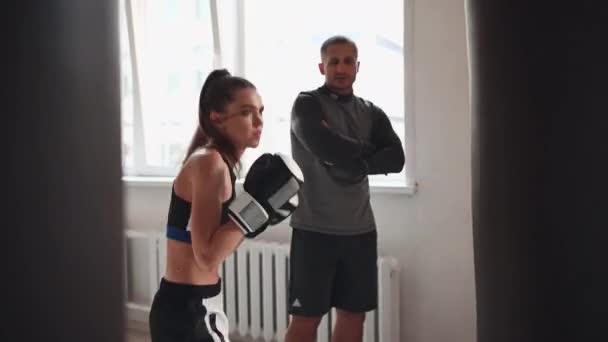 Un entraîneur strict regarde son étudiante kickboxer pratiquer un coup de poing sur un sac de boxe dans une salle d'entraînement spacieuse — Video