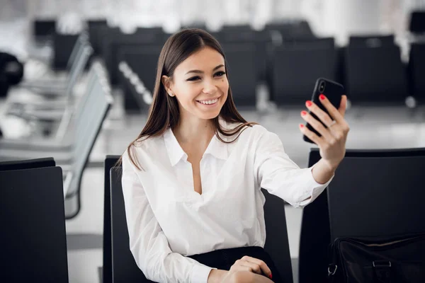 Joven mujer de negocios en el aeropuerto internacional, haciendo selfie con teléfono móvil y esperando su vuelo. Pasajera en la terminal de salida, en interiores. — Foto de Stock
