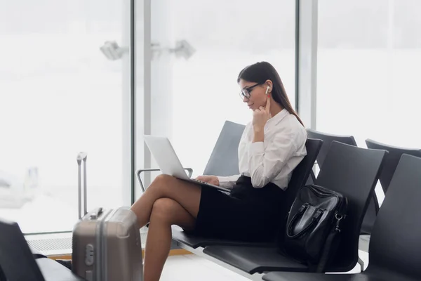 Молодая пассажирка аэропорта на смартфоне и ноутбуке сидит в терминальном зале в ожидании своего рейса. Концепция воздушных путешествий с молодой женщиной, сидящей с ручной клади — стоковое фото