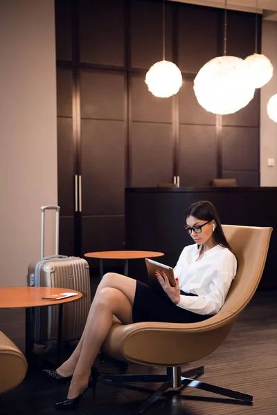 Empresaria sentada en la sala de negocios del aeropuerto, esperando el vuelo. Mujer sonriente con gafas sentada en el sofá en la sala de espera del aeropuerto. — Foto de Stock