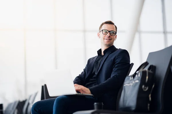 Man with suitcase sitting in airport waiting area while listening music using airpods — Stock Photo, Image