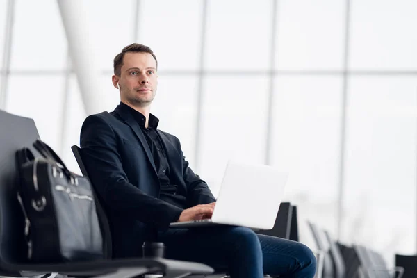 Mann mit Koffer sitzt im Flughafen-Wartebereich und hört Musik mit Airpods — Stockfoto