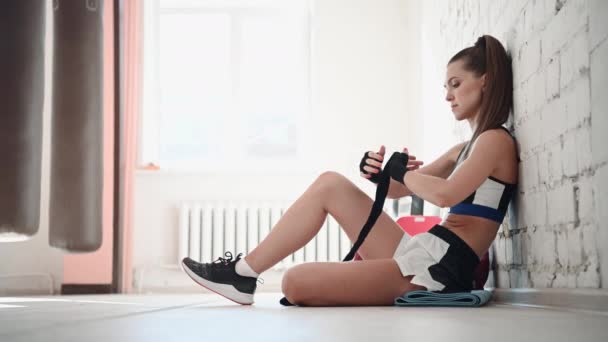 Ung kvinna som sitter på golvet och lindar in händerna med boxningslindor i fitnessklubben. Passar kvinnlig boxare som förbereder sig för träning. — Stockvideo
