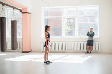 Geniş pencereli parlak bir spor salonunda genç bir bayan sporcu koçun gözetiminde ipe atlıyor.