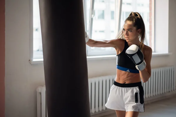 Una chica en ropa deportiva se dedica al boxeo y resuelve un golpe con la mano en un saco de boxeo — Foto de Stock