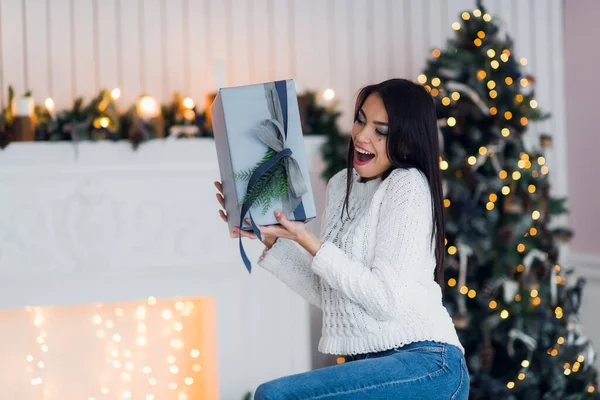 Happy Young Woman With Christmas Gift surprised and excited sitting at home against xmas decorations — Stock Photo, Image