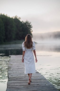 Back view of young woman in white dress standing alone on footbridge and staring at lake. Foggy chilly morning with a mist over water. clipart