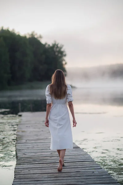 Rückenansicht einer jungen Frau in weißem Kleid, die allein auf einer Fußgängerbrücke steht und auf den See starrt. Nebel am kühlen Morgen mit Nebel über dem Wasser. — Stockfoto