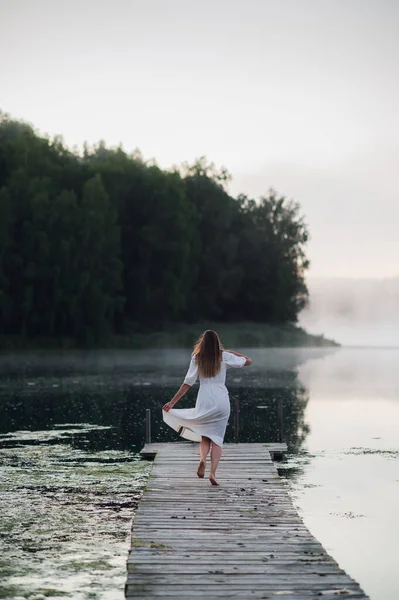 Frau im weißen hellen Kleid läuft über die Seebrücke am nebligen Morgensee. — Stockfoto