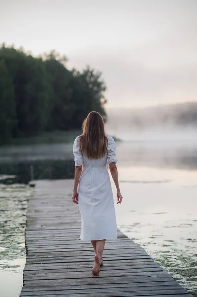 Vista posterior de la mujer joven en vestido blanco de pie solo en la pasarela y mirando al lago. Niebla mañana fría con una niebla sobre el agua. Imagen de stock