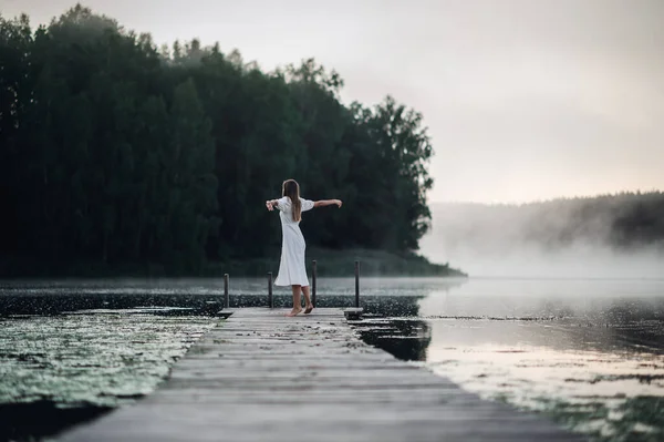 Vista posterior de la mujer joven en vestido blanco de pie solo en la pasarela y mirando al lago. Niebla mañana fría con una niebla sobre el agua. Fotos de stock