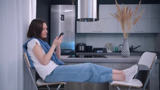 Kvinna vilar sittande i köket hålla smartphone välja varor på internet, göra mat leverans order on-line. Modern trådlös teknik förenklar livet, enkel användning av e-tjänster. — Stockvideo