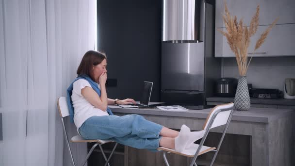 Evden uzak bir iş. Yorgun kadın esniyor, saatlerini kontrol ediyor işten sonra dizüstü bilgisayarını kapatıyor ve mola için mutfaktan çıkıyor. Karantina sırasında çalışan modern teknolojiyi kullanarak. — Stok video