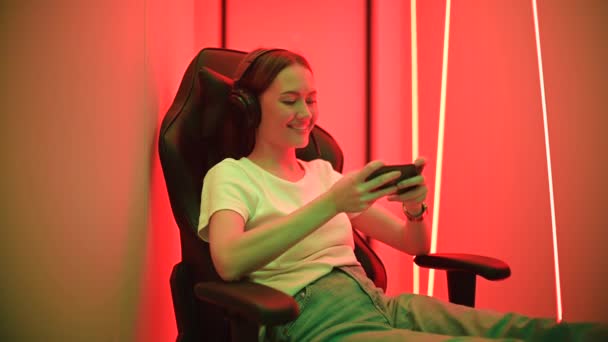 Веселая девушка играет в мобильную видеоигру, наслаждаясь классным приложением в гаджете, подсвеченном красным и зеленым неоновым цветом. — стоковое видео
