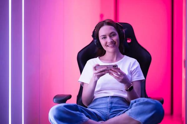 Молодая девушка играет в мобильную онлайн-игру на смартфоне, сидя на игровом стуле в красочном неоновом светлом помещении. — стоковое фото