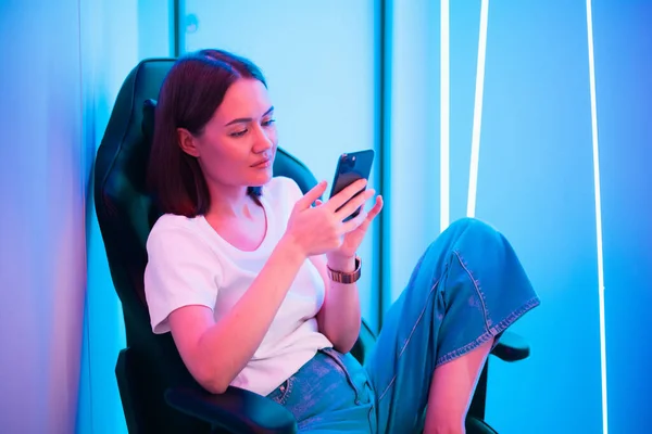 Νεαρή κυρία που κάθεται στην καρέκλα με το κινητό στα χέρια. Αναπαραγωγή online κινητό παιχνίδι ή περιήγηση στα κοινωνικά δίκτυα στηρίζεται σε ένα δωμάτιο φωτισμένο με νέον χρώμα. — Φωτογραφία Αρχείου