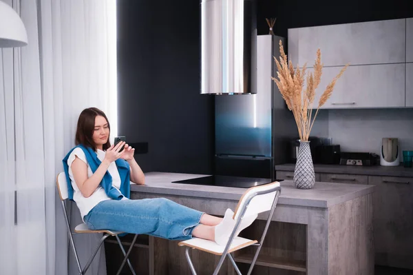Young beautiful woman using cell phone while sitting in the kitchen after coming back home from work. — Stock Photo, Image