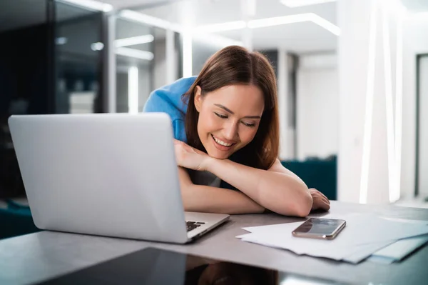 Mujer joven sonriente sentada en casa con el ordenador portátil, mirando a la pantalla del teléfono inteligente, esperando el mensaje de su novio o tienda de compras, pasar tiempo libre con el dispositivo móvil Imagen de archivo