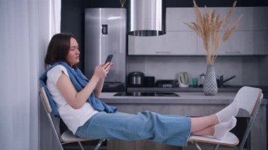 Genç ve güzel bir kadın işten eve döndükten sonra mutfakta otururken cep telefonu kullanıyor..