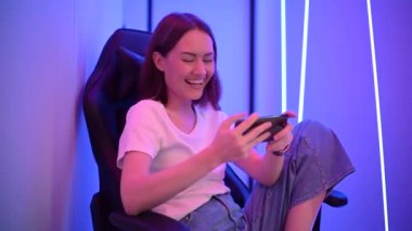 Cep telefonuyla oyun oynayan ve oyun sandalyesinde oturan bir kız. Renkli neon ışıklı oda.