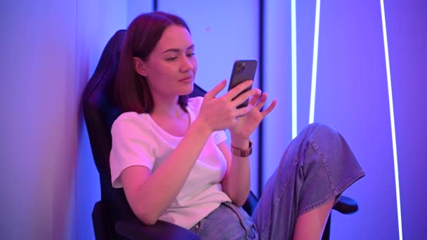 Młoda dama siedząca na krześle do gier z komórką w rękach. Gra online w gry mobilne lub przeglądanie serwisów społecznościowych odpoczywających w pokoju oświetlonym kolorem neonowym. — Wideo stockowe