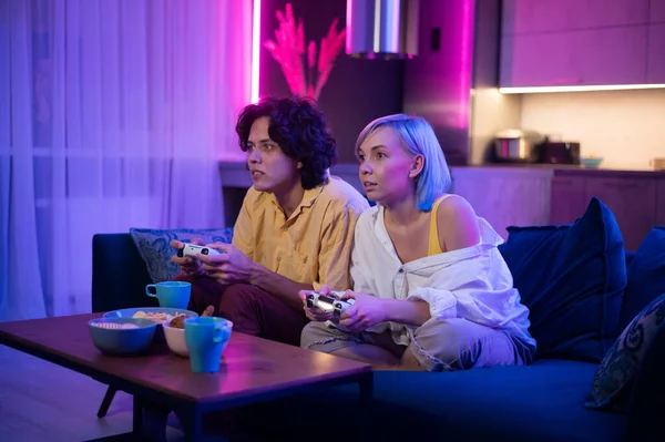Glückliche junge Leute, die Videospiele auf der Konsole spielen, während sie auf der Couch vor dem Fernseher sitzen. Millennial-Paar verbringt lustige Zeit zusammen zu Hause. Raum mit warmem Licht und Neonlicht. — Stockfoto