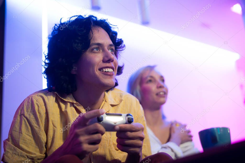 Young curly man and his girlfriend playing gaming console using joystick at home lit with neon pink and blue color.