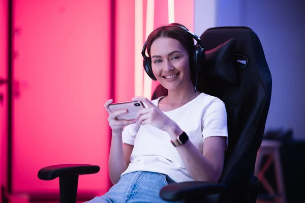 Young girl playing mobile online game on a smart phone while sitting on gaming chair in colorful neon light room. — Stock Photo, Image