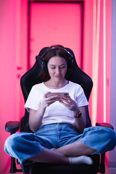 Cybersport-Gamer spielt Handyspiel auf dem Smartphone sitzend auf einem Gaming-Stuhl im neonfarbenen Raum. — Stockfoto