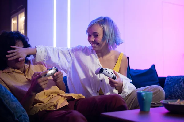 Pareja jugando videojuegos en la consola. Chica perder y no dejar que el chico jugar normalmente cerrando los ojos con su mano. Divertirse juntos en casa en cuarentena. — Foto de Stock