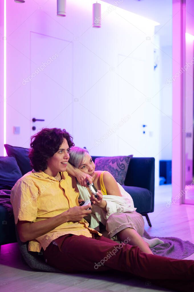 Happy boyfriend and girlfriend playing video games and holding joysticks at home. Millennial couple enjoing game while sitting on sofa and spending free time. Concept of relationship