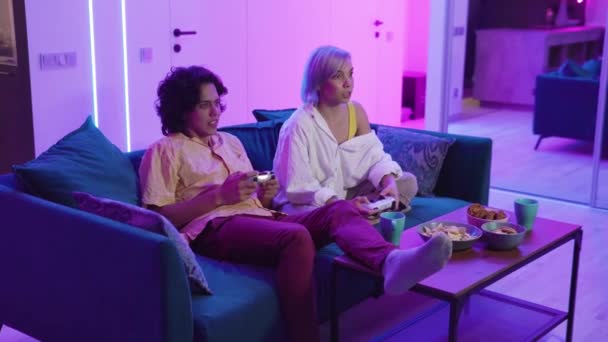 Šťastní mladí lidé hrají videohry na konzoli, zatímco sedí na gauči před televizí. Millennial pár tráví zábavný čas spolu doma. Pokoj s teplými a neonovými světly. — Stock video