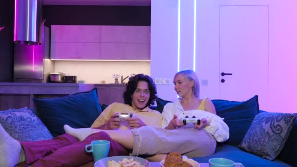 Szczęśliwy chłopak i dziewczyna śmieją się po graniu w gry wideo w domu, joysticks i przekąski na stole. Tysiącletnia para ciesząca się spędzaniem czasu razem. Pojęcie stosunku. — Wideo stockowe