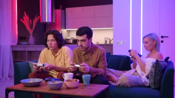 Due amici maschi entusiasti di giocare ai videogiochi a casa mentre una ragazza seduta con loro su un divano e utilizzando lo smartphone. — Video Stock